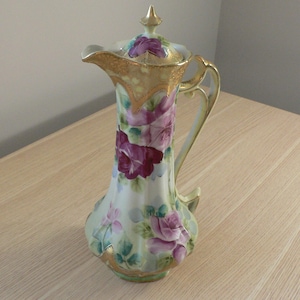 Może przedstawiać: Wysoki, zabytkowy dzbanek porcelanowy z pokrywką, z kwiatowym wzorem w odcieniach fioletu i różu. Dzbanek ma złoty uchwyt, obramowanie i akcenty. Tło jest jasnozielone. Wysokość ok. 25 cm.