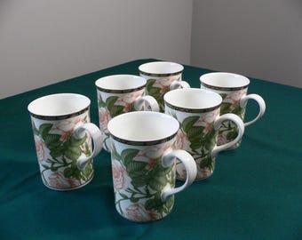 May include: Six white ceramic mugs with a floral design featuring pink roses and green leaves. The mugs have black trim around the top.