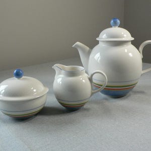 May include: A white ceramic tea set comprising a teapot, creamer, and sugar bowl. Each item features a blue knob and is adorned with horizontal stripes of blue, green, orange, and yellow. The teapot holds about 1.5 litres.