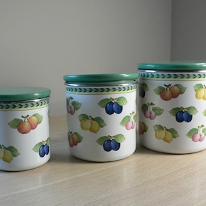 May include: A set of three white ceramic canisters with green lids and silver rims. Each canister features a fruit pattern with plums, peaches, and cherries. The canisters are of different sizes, suitable for kitchen storage.