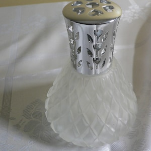 Könnte beinhalten: Ein klarer Glas-Ölbrenner mit strukturierter Oberfläche und einem silbernen Metalldeckel mit floralem Design.