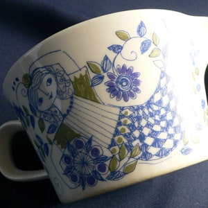 Może przedstawiać: Biały ceramiczny dzbanek do śmietanki z niebieskim i zielonym wzorem kwiatowym. Wzór przedstawia stylizowaną kobietę w sukience w kratę.