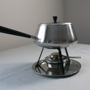 Può includere: Una pentola per fondue d'argento con un manico nero e un coperchio. La pentola è appoggiata su un supporto d'argento con un bruciatore nero e un vassoio d'argento.