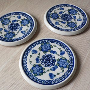 May include: Three round ceramic coasters with a white base and a blue floral design. The coasters feature a detailed pattern of blue flowers and leaves, with a decorative blue border. The coasters are arranged on a light-coloured wooden surface.