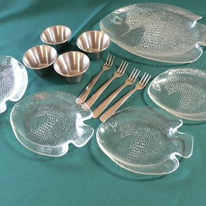 Peut inclure: Un ensemble de quatre plats de service en verre transparent en forme de poisson avec une surface texturée. Les plats sont disposés sur une nappe verte avec quatre petits bols en acier inoxydable et cinq fourchettes en argent.