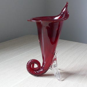 Puede incluir: Un jarrón de vidrio rojo con forma de cuerno con una base de vidrio transparente. El jarrón tiene un diseño de espiral y una abertura ancha.