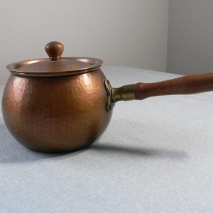 Puede incluir: Una cacerola de cobre martillada con un mango de madera y una tapa. La cacerola tiene un cuerpo redondeado y una textura martillada.