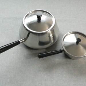 Pode incluir: Duas panelas de aço inoxidável com alças e tampas pretas. A panela maior tem capacidade para aproximadamente 1,4 litros, enquanto a menor tem cerca de 0,5 litros. Ambas têm um design clássico.