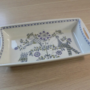 Könnte beinhalten: Rechteckige Servierplatte aus Keramik mit Griffen. Die weiße Schale hat ein blaues und violettes Blumen- und Figuren-Design. Der Rand und die Griffe sind mit einem blauen Muster verziert. Ein dekoratives Stück zum Servieren.