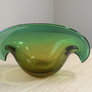 May include: A green glass bowl with a distinctive, curved design. The bowl features a wide, open top with flared edges that curve outwards. The glass has a gradient of colours, transitioning from a deep green to a lighter, yellowish-green hue.