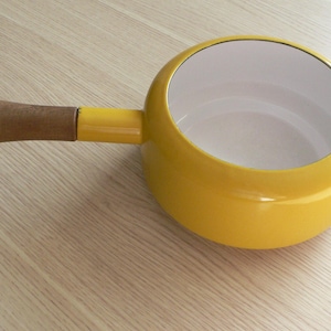 Può includere: Una casseruola gialla brillante con interno bianco e manico in legno. La casseruola è rotonda con un lungo manico. Il manico è in legno. Appoggiata su una superficie a venature di legno chiaro.