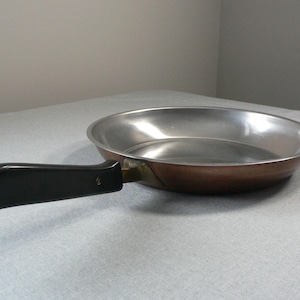 Puede incluir: Una sartén de color cobre con interior de acero inoxidable y mango negro. La sartén tiene forma redonda y está diseñada para cocinar. El mango está sujeto con remaches de latón.