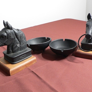 Puede incluir: Dos ceniceros de hierro fundido negro en forma de cabeza de caballo con bases de madera. Cada cenicero tiene un cuenco redondo y una cabeza de caballo con un anillo en la boca. Las cabezas de caballo están sobre una base de madera.