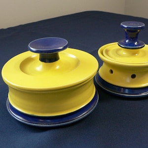 May include: Two yellow ceramic containers with blue lids and blue trim. The smaller container has holes in the bottom.
