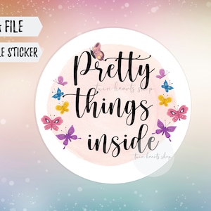 May include: A circular sticker design featuring the black text Pretty things inside centered over a soft pink watercolor circle. The design is surrounded by small, colorful butterflies in shades of pink, purple, yellow, and blue. Two white banners at the top left read PNG FILE and PRINTABLE STICKER.