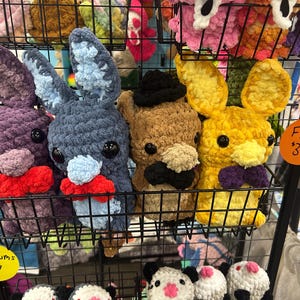 May include: A display of handmade crochet bunny plushies in various colors, including purple, blue, brown, and yellow. Each bunny has large ears, black eyes, and a bow tie or hat. The sign says "FNAF $35".