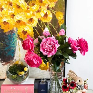 Peut inclure: Nature morte vibrante avec un bouquet de roses et de pivoines roses dans un vase en verre transparent. Une peinture de fleurs jaunes dans un vase bleu est en arrière-plan. Un petit terrarium et un lapin décoratif sont également présents.