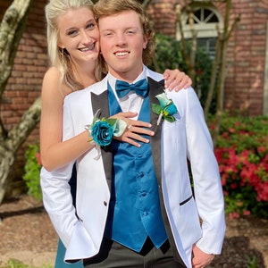 May include: A white tuxedo jacket with black lapels, a teal blue vest, a matching teal blue bow tie, and a teal rose boutonniere. The ensemble is paired with black trousers, creating a formal look with vibrant teal accents and crisp white fabric.