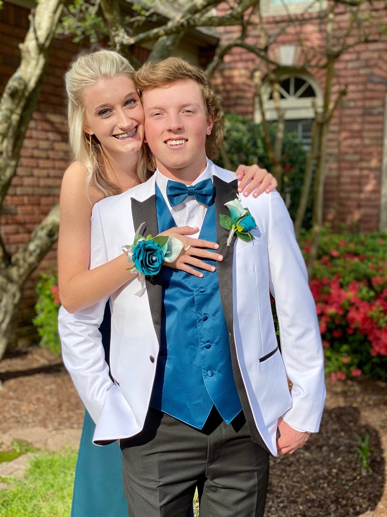 May include: A white tuxedo jacket with black lapels, a teal blue vest, a matching teal blue bow tie, and a teal rose boutonniere. The ensemble is paired with black trousers, creating a formal look with vibrant teal accents and crisp white fabric.