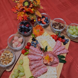 May include: A wooden charcuterie board featuring a selection of snacks, such as salami, cheese, crackers, grapes, strawberries, and blueberries. The board is adorned with a flower and foliage.
