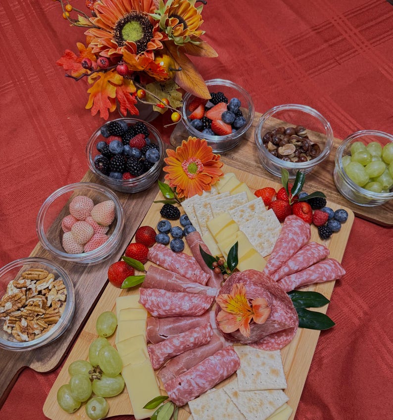 May include: A wooden charcuterie board featuring a selection of snacks, such as salami, cheese, crackers, grapes, strawberries, and blueberries. The board is adorned with a flower and foliage.