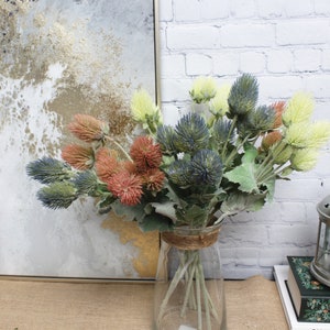 Puede incluir: Un ramo de flores de cardo artificiales en un jarr&oacute;n de vidrio transparente. El ramo presenta flores de cardo azules, verdes y rosas con hojas verdes.