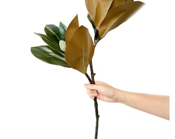 May include: A single stem of artificial magnolia leaves in shades of brown and green. The leaves are large and have a glossy finish.