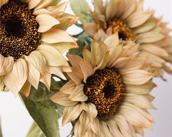 Pode incluir: Close-up de vários girassóis artificiais com pétalas em camadas amarelo-claras e centros castanhos escuros salpicados de amarelo. Os girassóis têm folhas e caules verdes. As flores estão dispostas em um buquê.