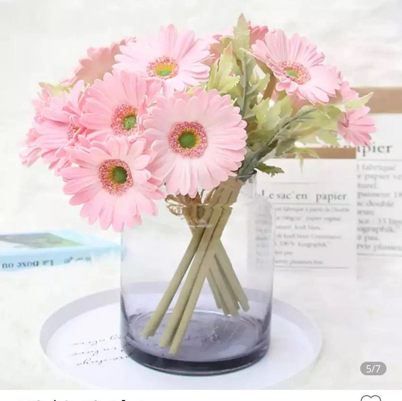 May include: A bouquet of pink artificial gerbera daisies in a clear glass vase with a grey tint. The vase is sitting on a white surface.