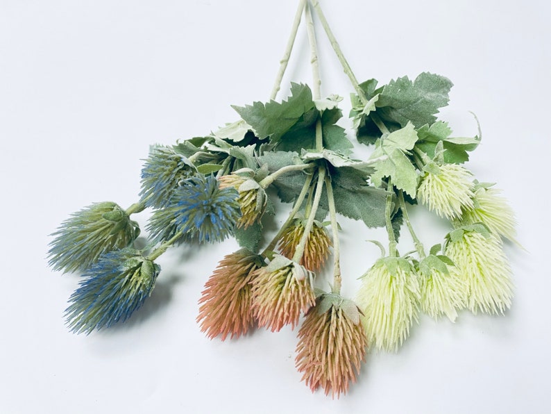 Puede incluir: Un ramo de flores artificiales con flores azules, rojas y crema. Las flores tienen una textura espinosa y est&aacute;n dispuestas en tallos verdes.