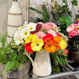 May include: A bouquet of colourful gerbera daisies in a white ceramic jug. The flowers are arranged in a variety of colours, including pink, red, yellow, and orange. The jug is sitting on a table with a green plant in a wooden pot.