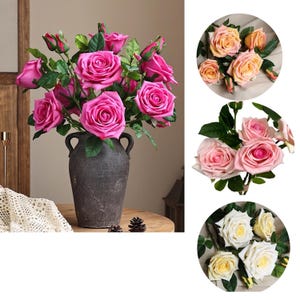 May include: A bouquet of artificial pink roses in a dark gray vase. The roses are arranged in a cluster with green leaves. The vase has two handles on the sides. The bouquet is on a wooden table.
