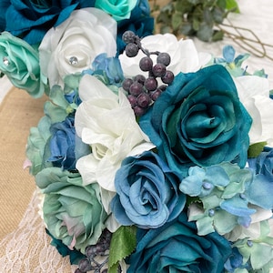 May include: A floral bouquet featuring artificial roses in shades of deep teal, dusty blue, and white, accented with light mint green petals and clusters of small dark purple berries. The arrangement includes various fabric flower heads and greenery.