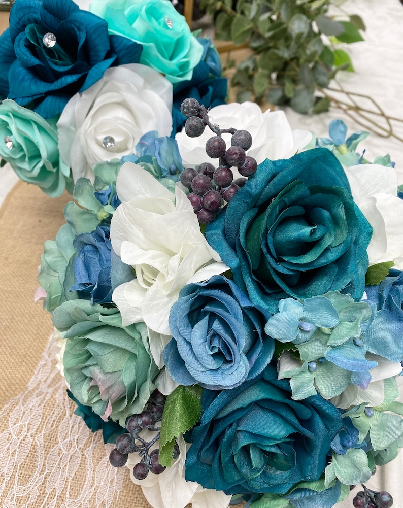May include: A floral bouquet featuring artificial roses in shades of deep teal, dusty blue, and white, accented with light mint green petals and clusters of small dark purple berries. The arrangement includes various fabric flower heads and greenery.