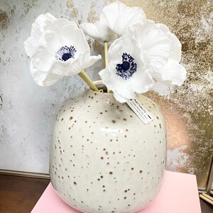 May include: White paper anemones with dark blue centers in a speckled white ceramic vase. The vase is sitting on a pink surface.