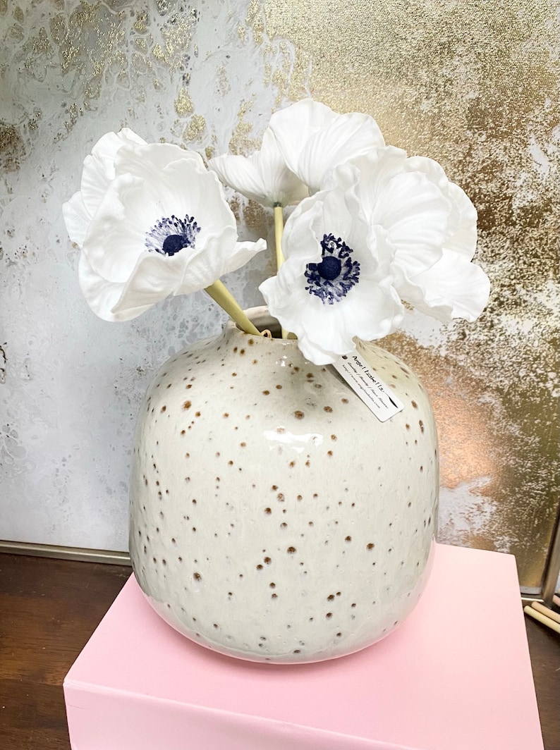 May include: White paper anemones with dark blue centers in a speckled white ceramic vase. The vase is sitting on a pink surface.