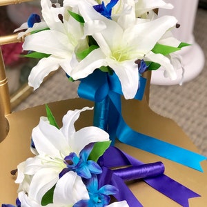 May include: Two white lily bouquets with blue orchid accents. The larger bouquet has a teal ribbon, while the smaller bouquet has a purple ribbon.