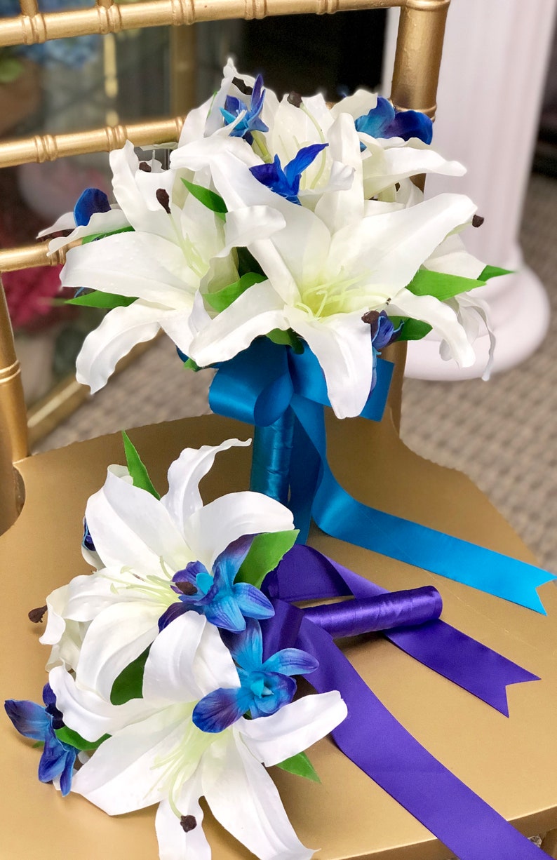 May include: Two white lily bouquets with blue orchid accents. The larger bouquet has a teal ribbon, while the smaller bouquet has a purple ribbon.