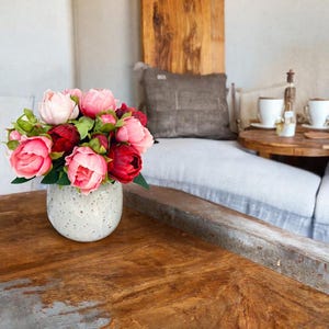 May include: A bouquet of pink and red peonies in a white speckled ceramic vase sits on a wooden table.