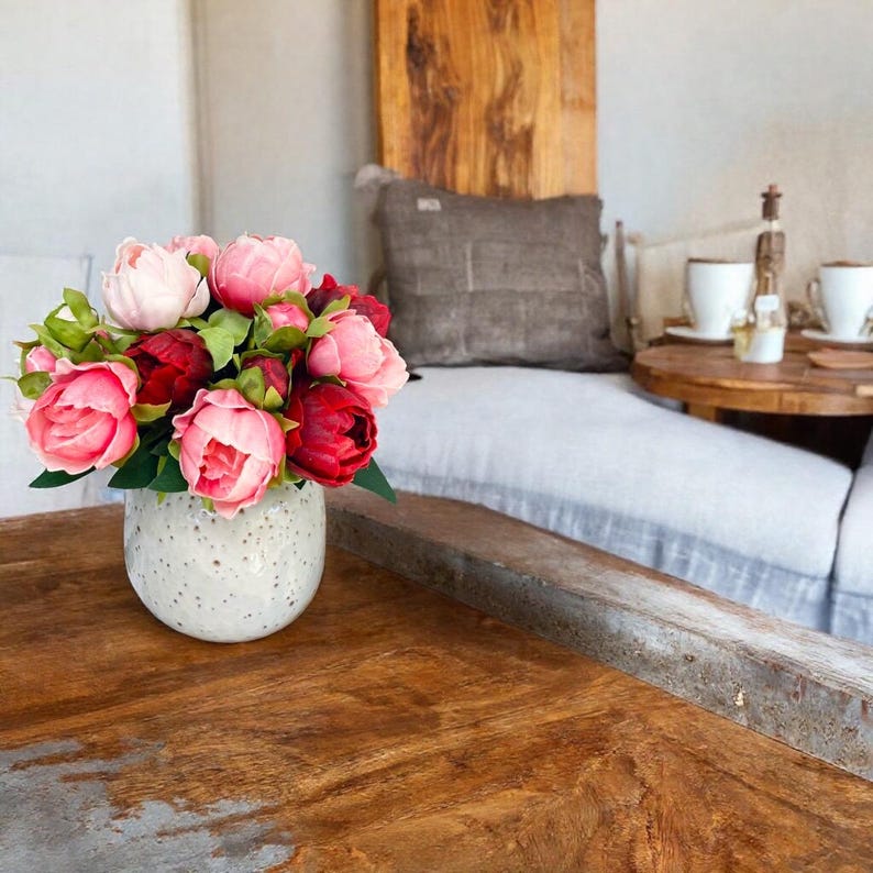 May include: A bouquet of pink and red peonies in a white speckled ceramic vase sits on a wooden table.
