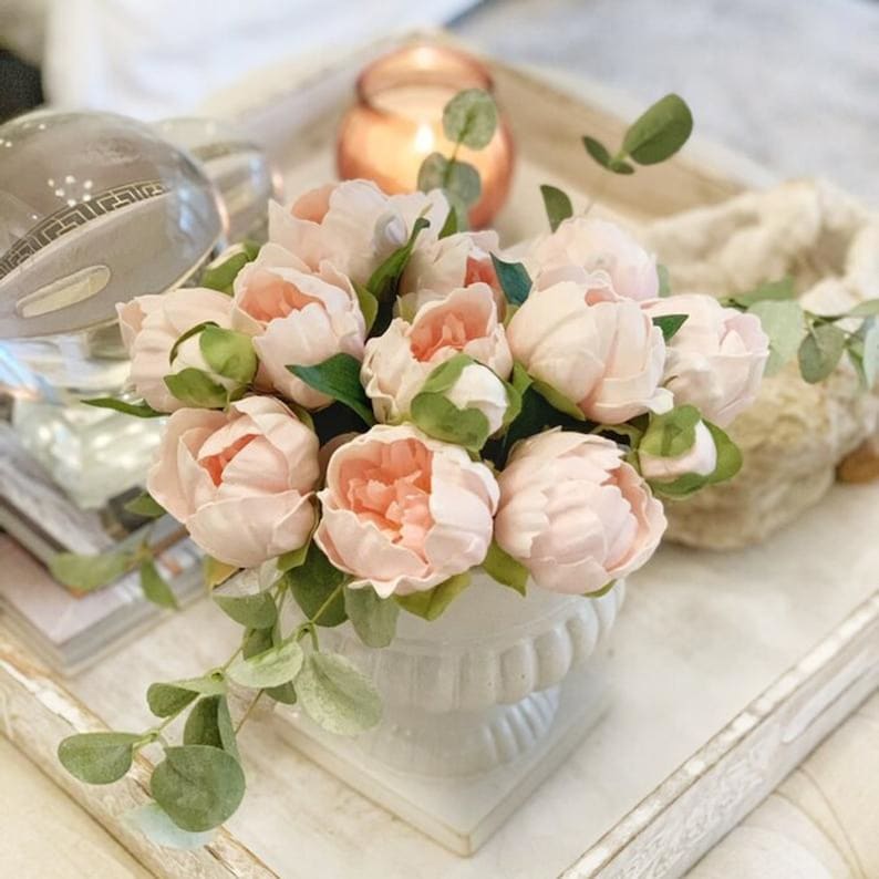 May include: A bouquet of pale pink peonies arranged in a white ceramic vase with green eucalyptus sprigs. The flowers are on a white tray with a decorative edge.