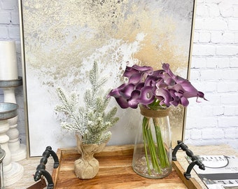 May include: A wooden tray with a faux snow-covered pine sprig and a glass vase filled with purple calla lilies. The tray is in front of a canvas with a gold and white abstract design.