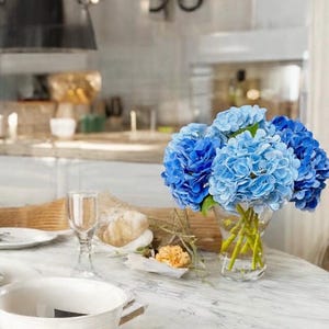 Pode incluir: Um buquê de hortênsias azuis em um vaso de vidro transparente sobre uma mesa de mármore branco. O vaso está sobre uma toalha de mesa branca com uma tigela branca e um copo.