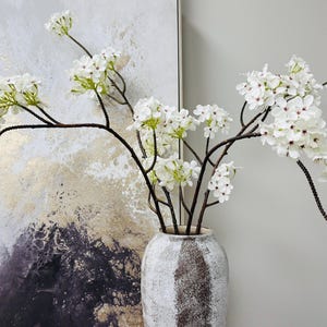 May include: A vase of white flowers with dark brown stems. The flowers have small petals with red centers. The vase is off-white and textured. The arrangement is in front of a painting with gold, white, and black tones.
