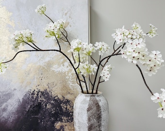 Puede incluir: Un arreglo decorativo de flores blancas con tallos marrones oscuros en un jarrón texturizado blanquecino. Las flores tienen pequeños pétalos delicados con centros rojos. El jarrón está frente a una pintura con tonos dorados, blancos y negros.