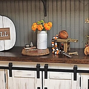 May include: White wooden pumpkin with the words "Hello Fall" on it, a white mason jar with orange flowers, a wooden stool with a brown acorn on top, a tiered tray with fall decorations, and a white cabinet with black hardware.