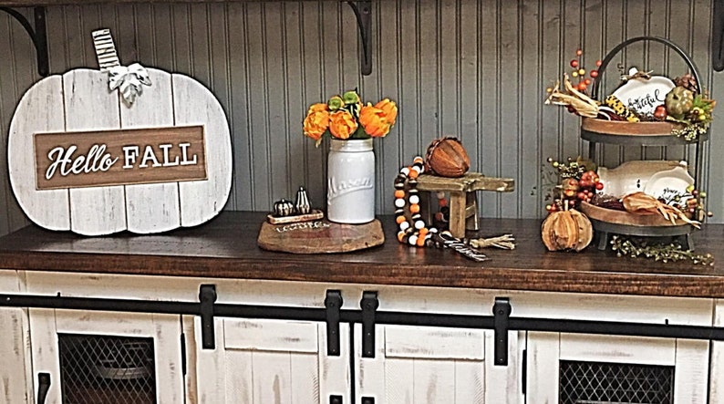 May include: White wooden pumpkin with the words "Hello Fall" on it, a white mason jar with orange flowers, a wooden stool with a brown acorn on top, a tiered tray with fall decorations, and a white cabinet with black hardware.