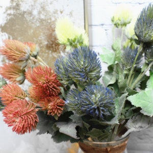 Puede incluir: Un ramo de flores artificiales con flores azules, rojas y amarillas que se asemejan a cardos. Las flores est&aacute;n dispuestas en un jarr&oacute;n.
