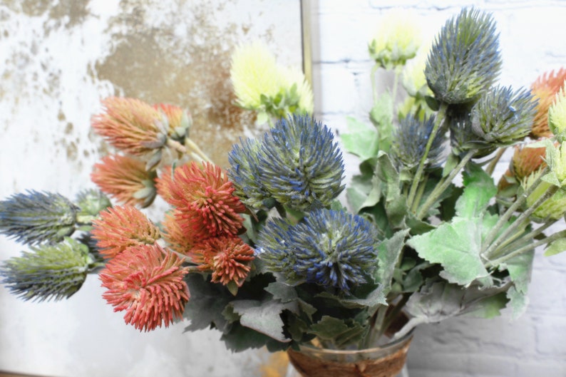 Puede incluir: Un ramo de flores artificiales con flores azules, rojas y amarillas que se asemejan a cardos. Las flores est&aacute;n dispuestas en un jarr&oacute;n.