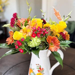 May include: A bouquet of yellow, orange and red flowers in a white jug with a pumpkin design. The flowers are arranged in a vase with a white background.